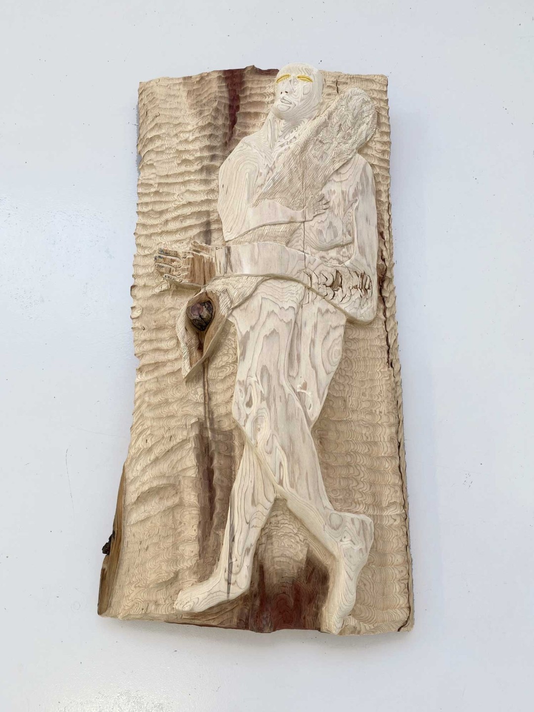 Nous voyons un bois de séquoia de 150 centimètres de haut pour 75 centimètres de large. Dans le bois est scupté un figure humaine portant un rondin de bois.