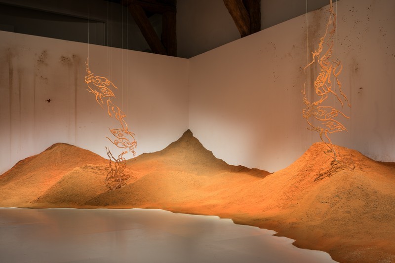 Large monticule et de sable mélangé à des épices au dessus duquel est suspendues deux sculptures en acier découpé, colorées avec des épices et rappelant la calligraphie arabe