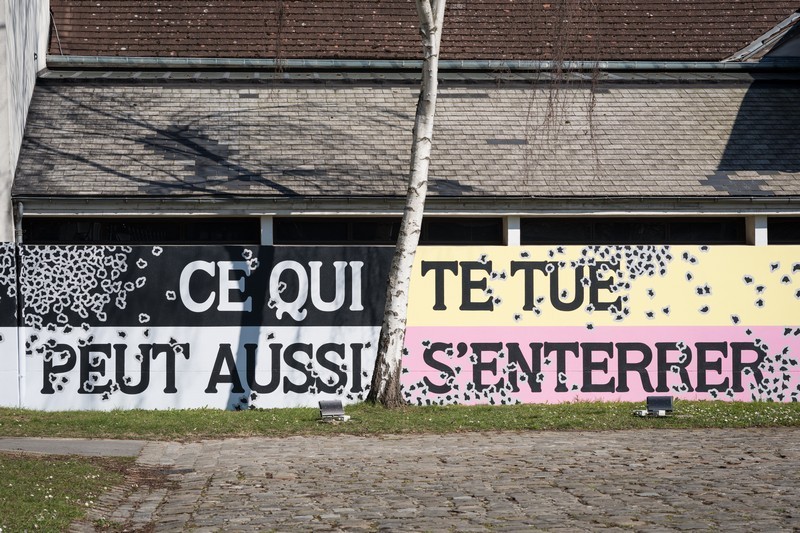 Fresque murale extérieure avec le texte « Ce qui te tue peut aussi s'enterrer », peint en grandes lettres majuscules blanches et noires sur des bandes de couleurs noir, jaune pâle et rose, parsemées de formes tachetées