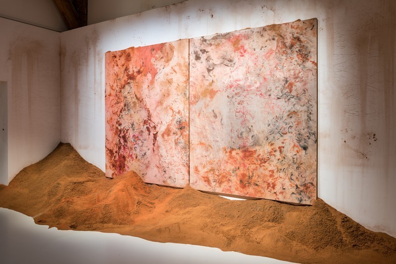 Deux toiles abstraites au fusain et aux épices accrochées l'une à côté de l'autre, bord à bord au dessous d'un monticule de sable coloré aux épices