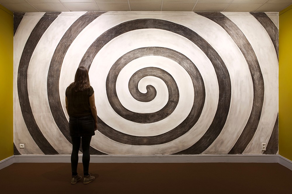 Une personne de dos regarde une spirale en noir et blanc qui occupe un mur complet