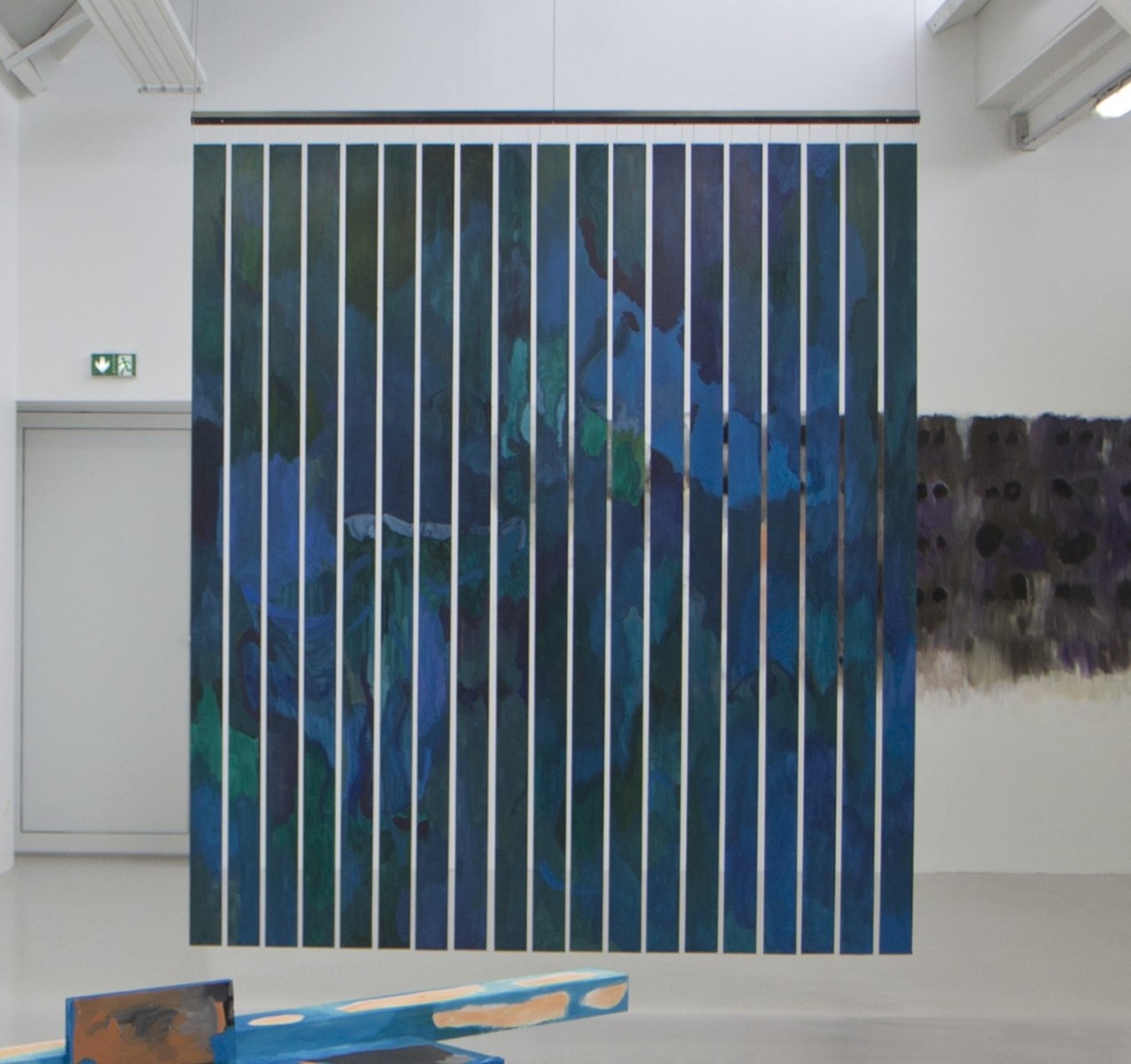 L'oeuvre est une peinture acrylique sur des lames de bois disposées verticalement, alignées horizontalement, et suspendues. La peinture prend des formes colorées sur des tons bleus et verts.