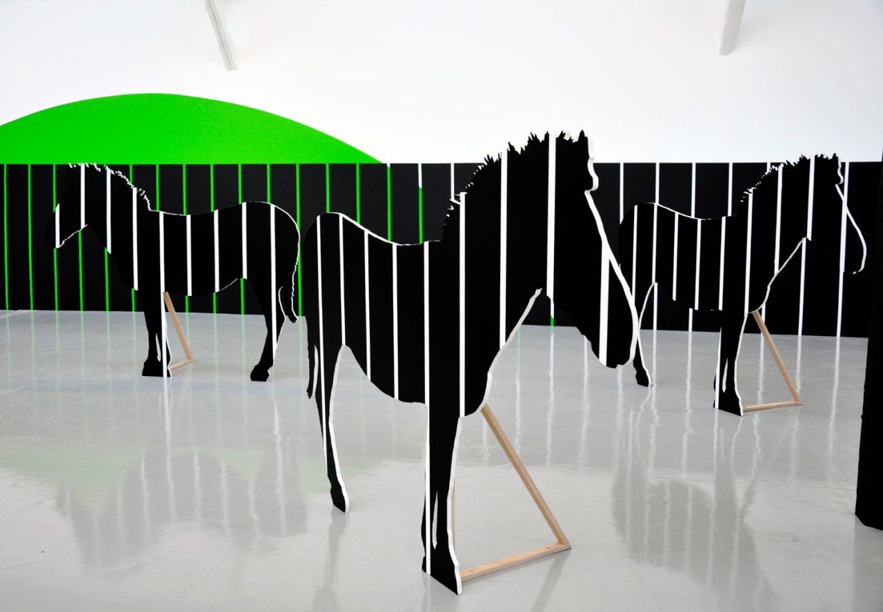 Vue de l'exposition. Plusieurs chevaux noir avec des rayures blanches en carton se tiennent debout dans la salle d'exposition