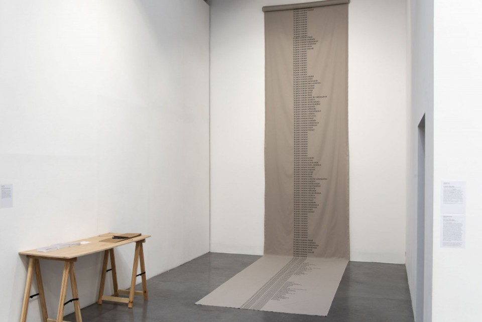 Un long tissu en coton beige se déroule depuis le plafond. Un poème est imprimé. Chaque phrase commence par "le pain comme". Une table accueille des cartes postales pour que les gens complètent la phrase. Il y a également un cahier pour qu'ils puissent écrire la suite du poème.