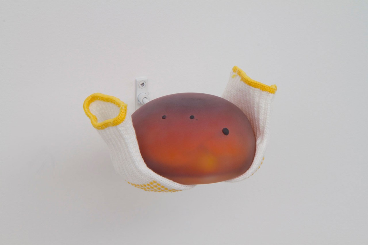 L'oeuvre est constituée d'une forme ronde en verre soufflé accrochée au mur avec des gants en tissus et plastiques collés sous celle-ci, comme si les gants la portaient.