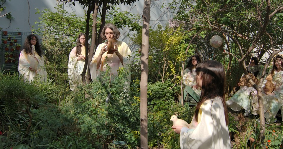 Image extraite de la vidéo Le Cantique des oiseaux de Katia Kameli. Des musiciennes jouent de la flute dans un jardin arborée. Elles sont vetues de robes amples aux couleurs pastels.